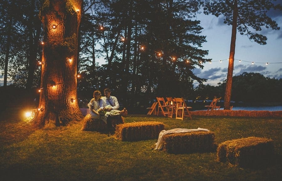 The bride and groom sit on hay bails and talk to each other as the sun goes down at Brook farm