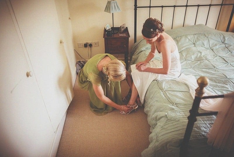 A bridesmaid fastens the buckle of the brides shoe as the bride sits on her mothers bed