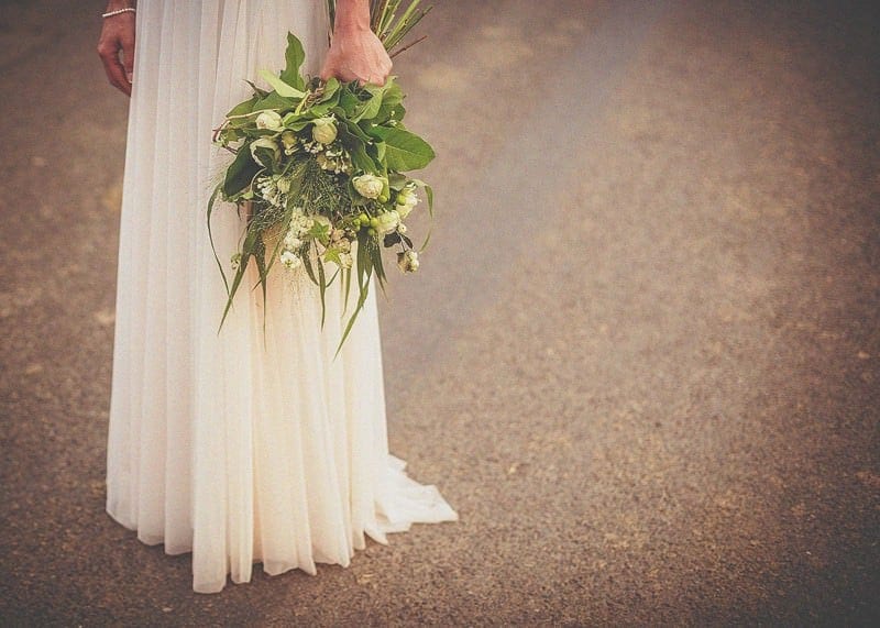 The bride stands in the middle of the road holding her bouquet next to her side