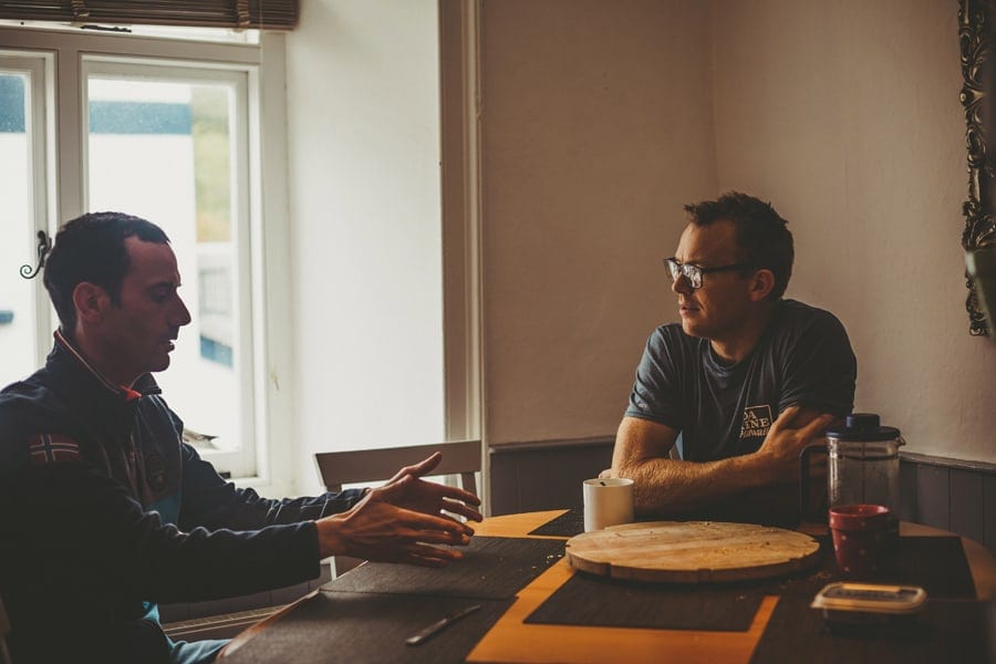 Two men sit and talk at a wooden table next to a window and drink coffee