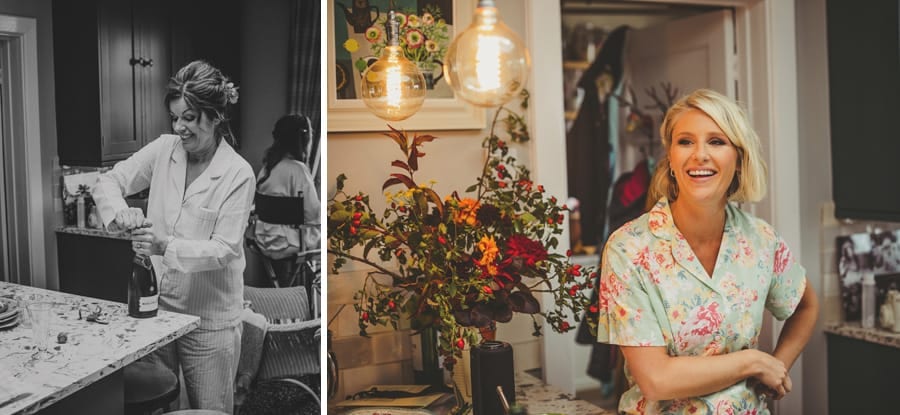 A bridesmaid stands in the kitchen and smiles as the brides mother attempts to open a bottle of champagne