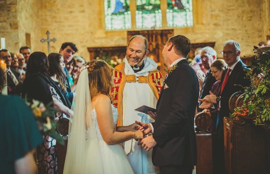 The vicar smiles at the bride and both the bride and groom laugh at the vicar in the church