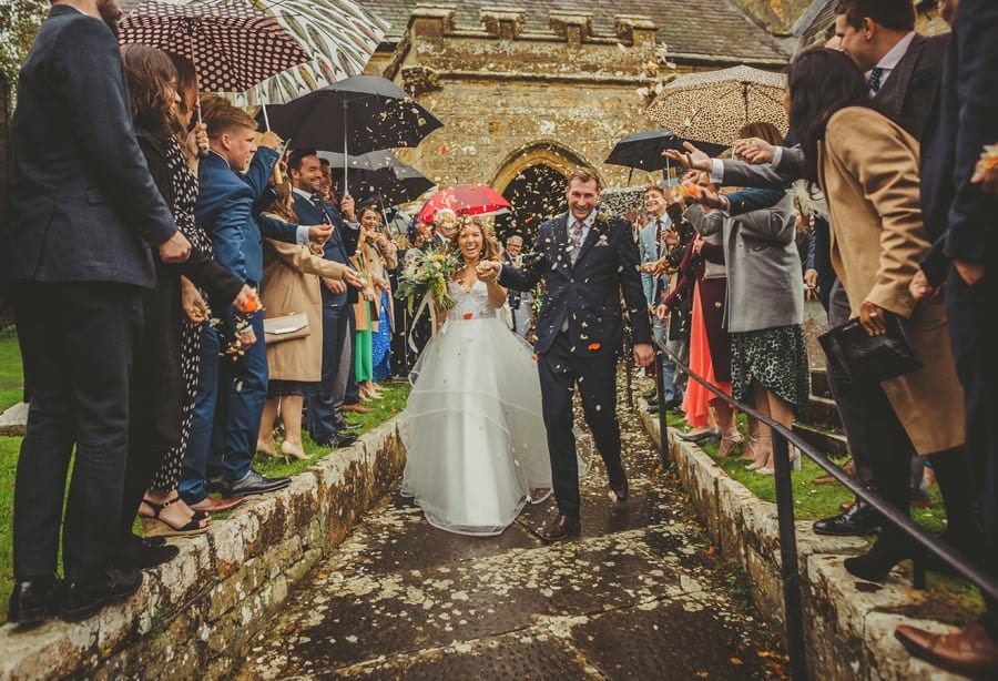 The bride and groom walk down the path of the church as wedding guests throw confetti in the air
