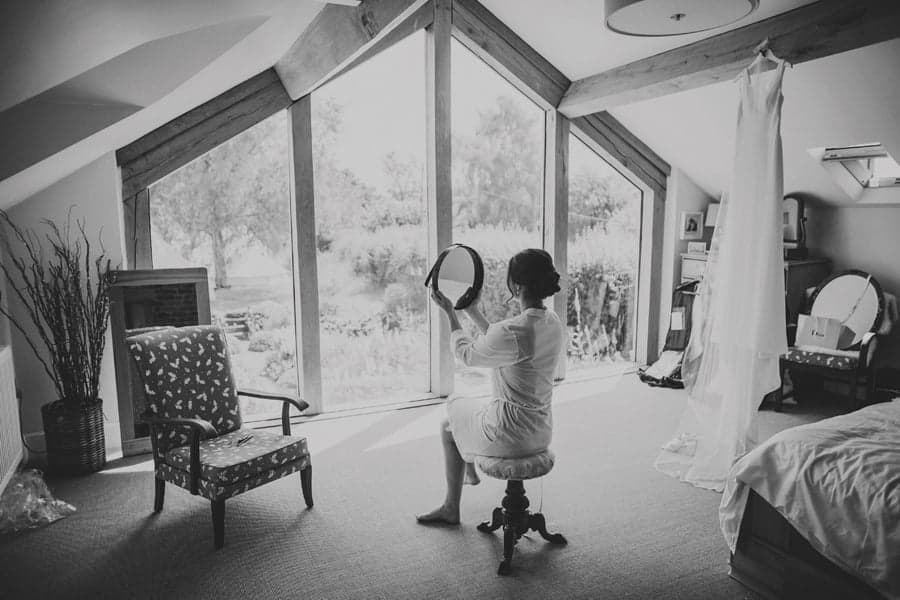 The bride holds up a mirror and looks at herself sat on a stool in the bedroom at Mill farm