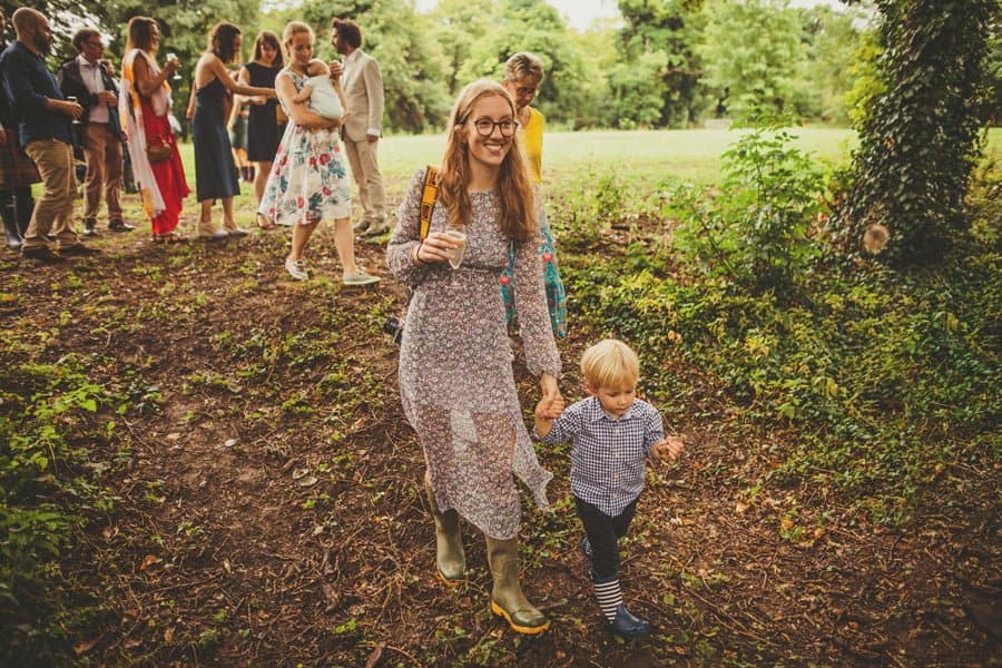 A wedding guest walks into the woods holding a glass of champagne with her little boy