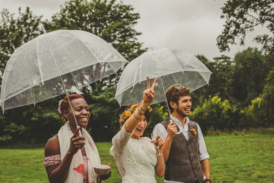 The bride, groom and one of her bridesmaids stand under umbrellas in the school field watching the games