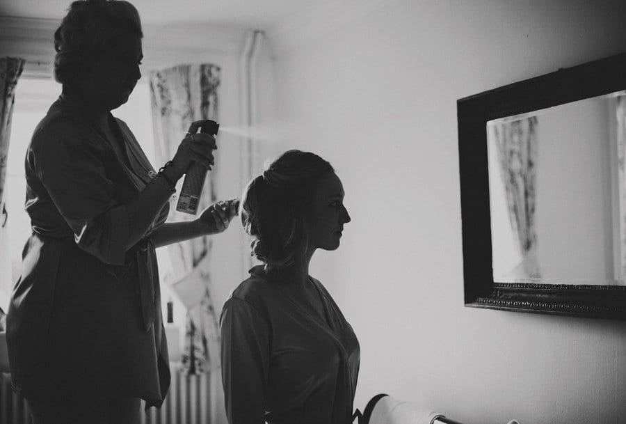 A bridesmaid stands on a chair and sprays hairspray on a friends hair as she looks into the mirror