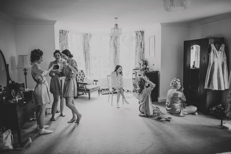 The bride sits on a chair in the middle of the room and watches the bridesmaids getting ready