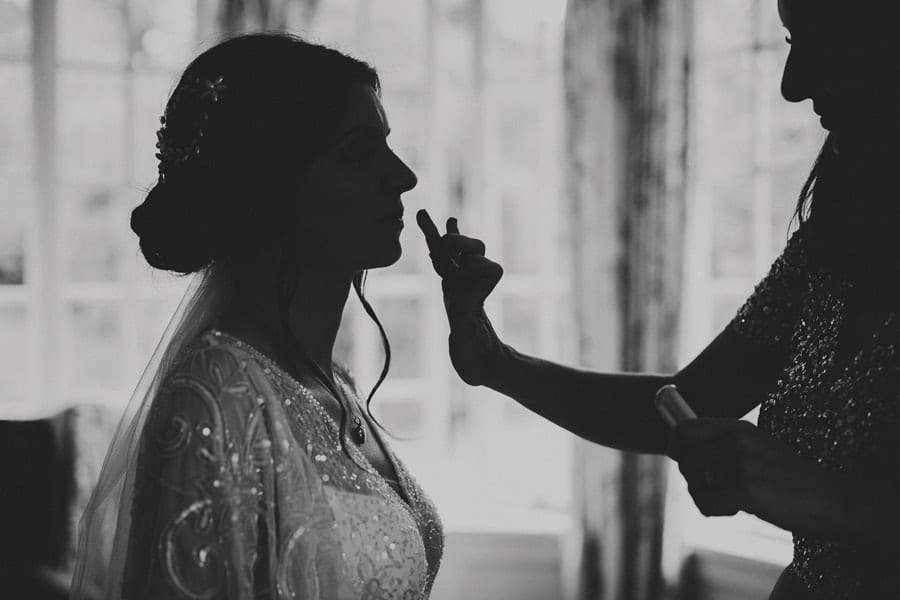 A bridesmaid places lip balm onto the lips of the bride in the main bedroom at Barley Wood house