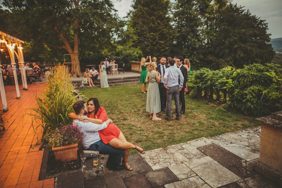 A lady sits on a mans legs at the side of Barley Wood house in the evening