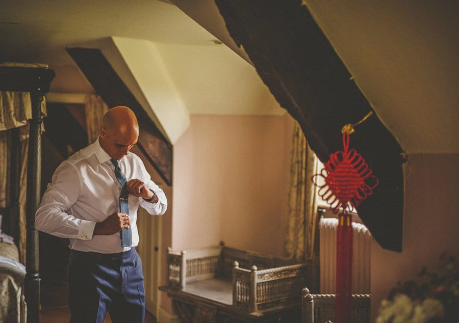 The groom looks down and straightens his tie in a bedroom at Abbey House Gardens