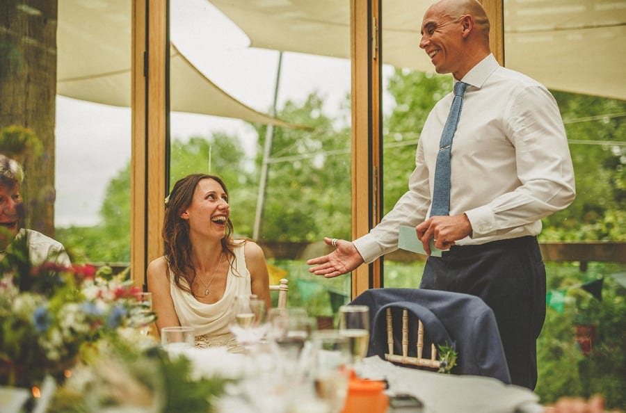 The groom stands up and delivers his speech as the bride looks on and laughs