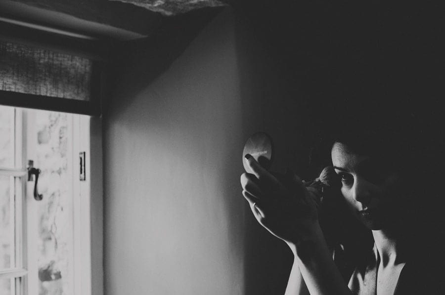 A bridesmaid applies makeup to her face next to a window in one of the bedrooms of the cottage