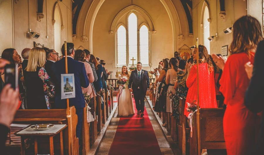 The bride and groom walk down the aisle of the Church together as wedding guests stand up and clap their hands