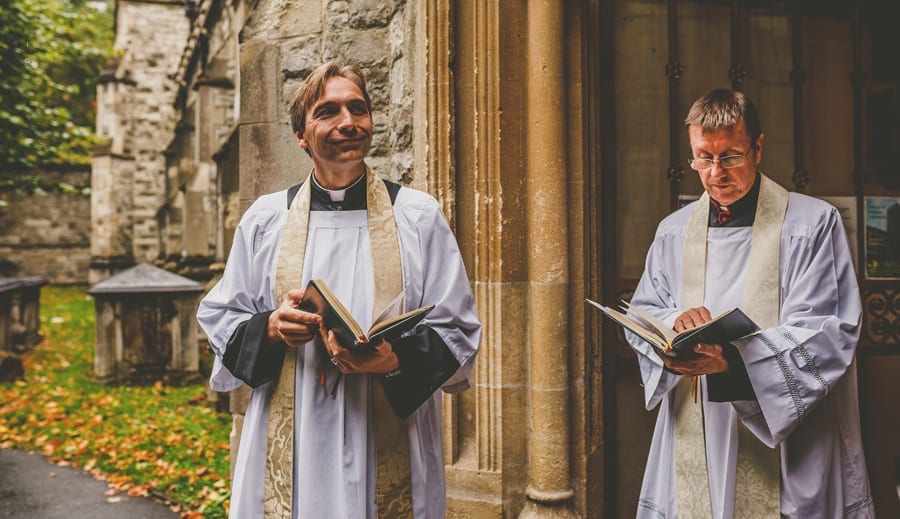 The vicar holds a bible and waits for the bridal party outside the Church at Fulham Palace