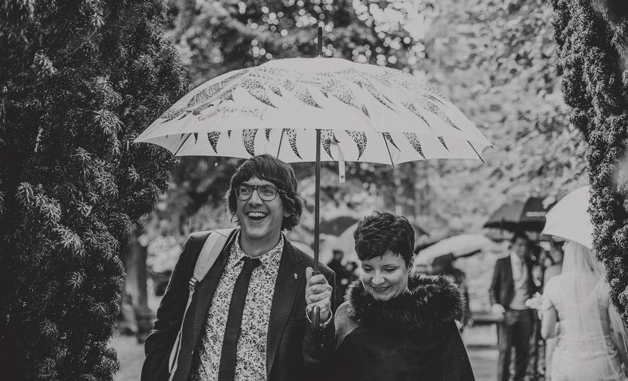 A man holding an umbrella and a lady at his side smile as they walk through the gardens of the Church