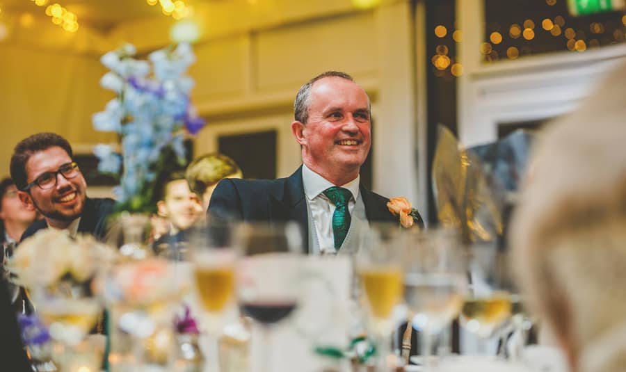 The grooms father laughs as he sits at the table and listens to the best mans speech
