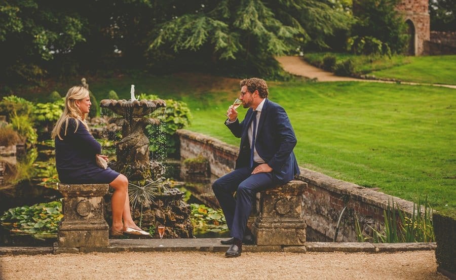A wedding guest sits on a wall and drinks champagne