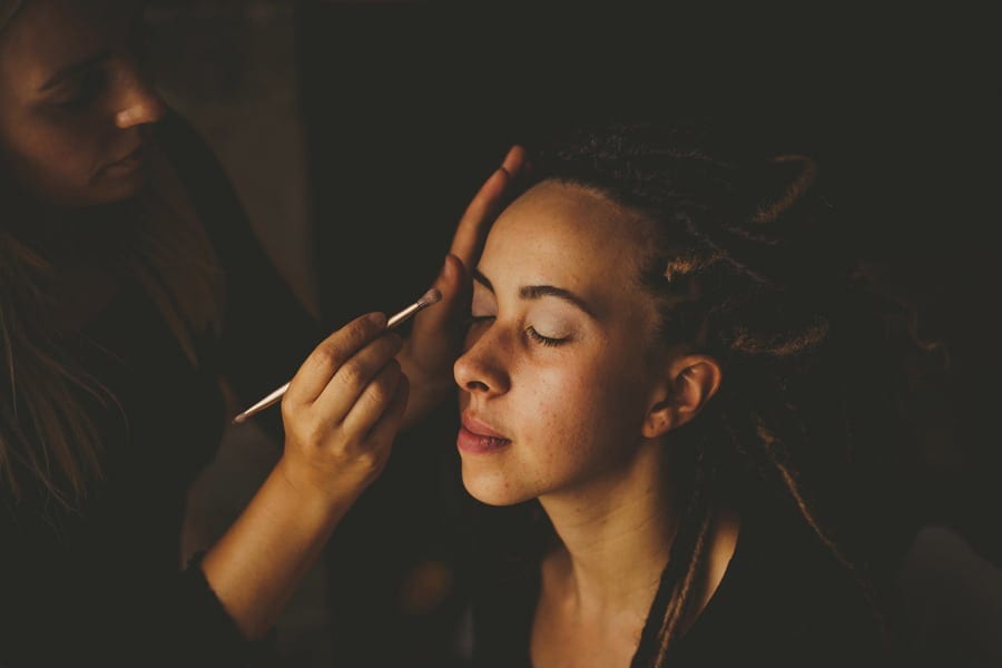 The bride sits next to a window and has her make up applied onto her eyebrows
