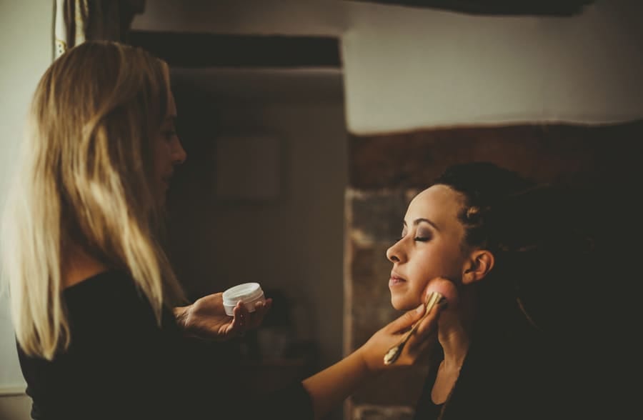 The makeup artist applies make up on the brides face next to a window in the brides bedroom