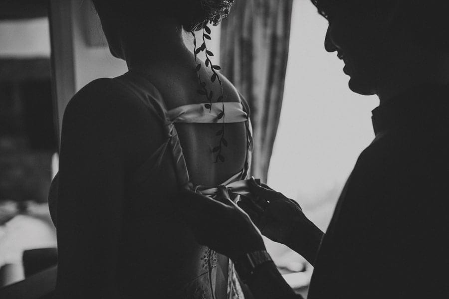 The brides friend ties the back of a dress next to a window
