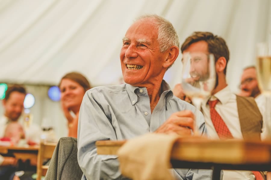 A wedding guest sits at the table in the marquee and smiles as he listens to the groom's speech
