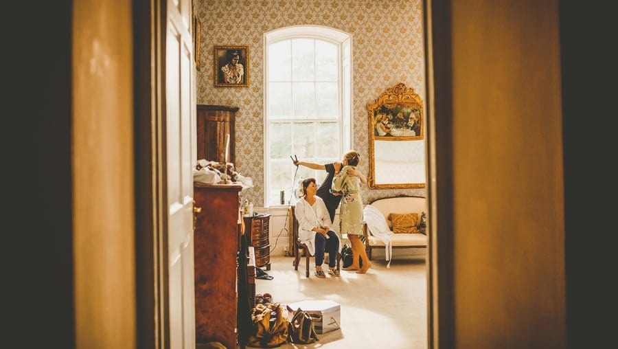 The brides mother hugs the make up artist as a bridesmaid sits on a chair next to a large window at Kings Weston House
