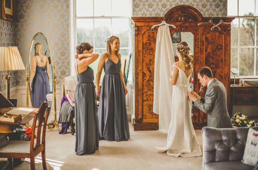 A wedding guest ties the back of the brides wedding dress as the bridesmaids talk to the bride