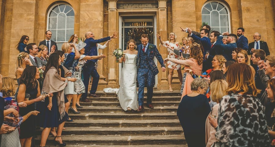 The bride and groom walk down the steps outside Kings Weston House as wedding guest throw confetti in the air