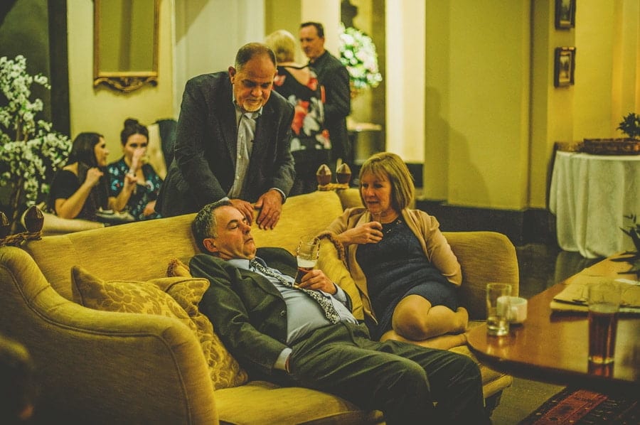A man stands over a wedding guest who is asleep on the couch as a lady looks on