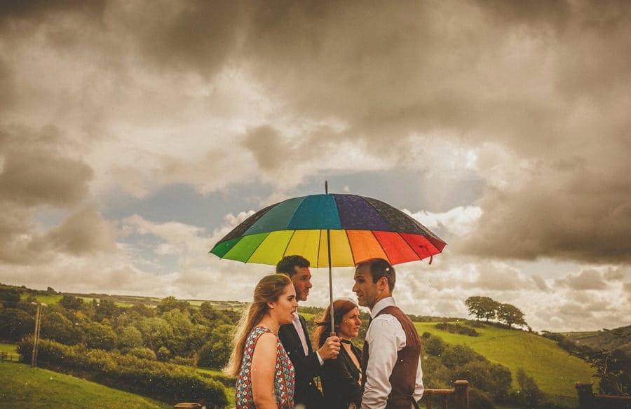 Little Quarme wedding photographer 0020 | somerset-wedding-photographer Wedding guests wait under a multi coloured umbrella in a field at Little Quarme Cottages