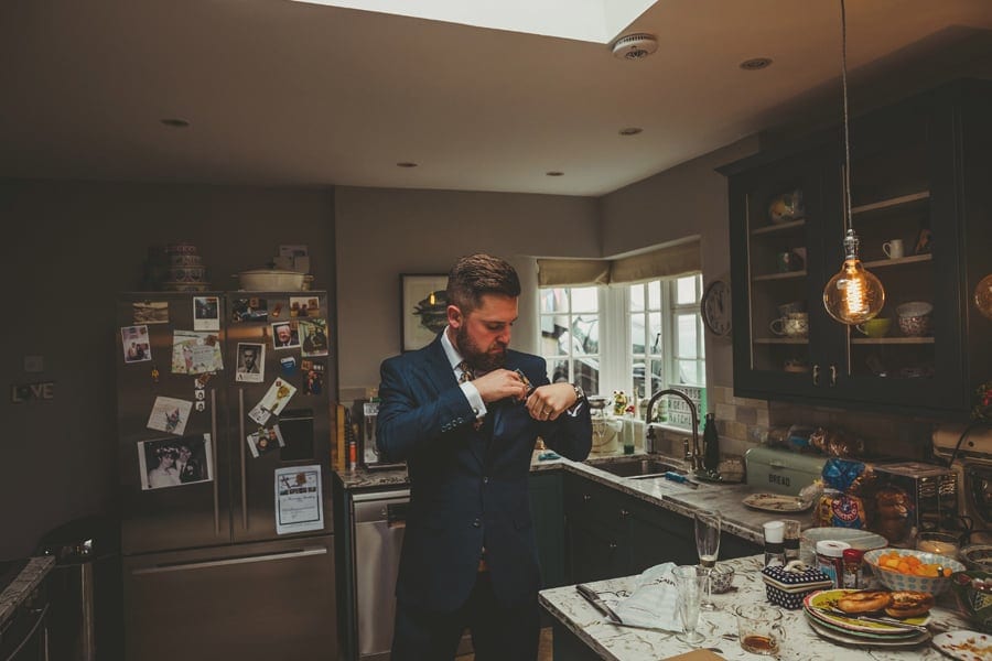 A man places a handkerchief into his suit pocket in the kitchen