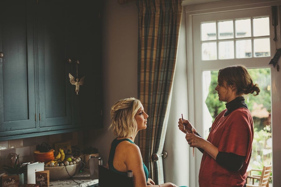A bridesmaid sits next to a large window as the make up artist looks at her