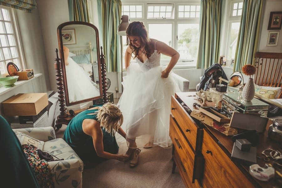 A bridesmaid helps the bride put on her new shoes in her parents bedroom