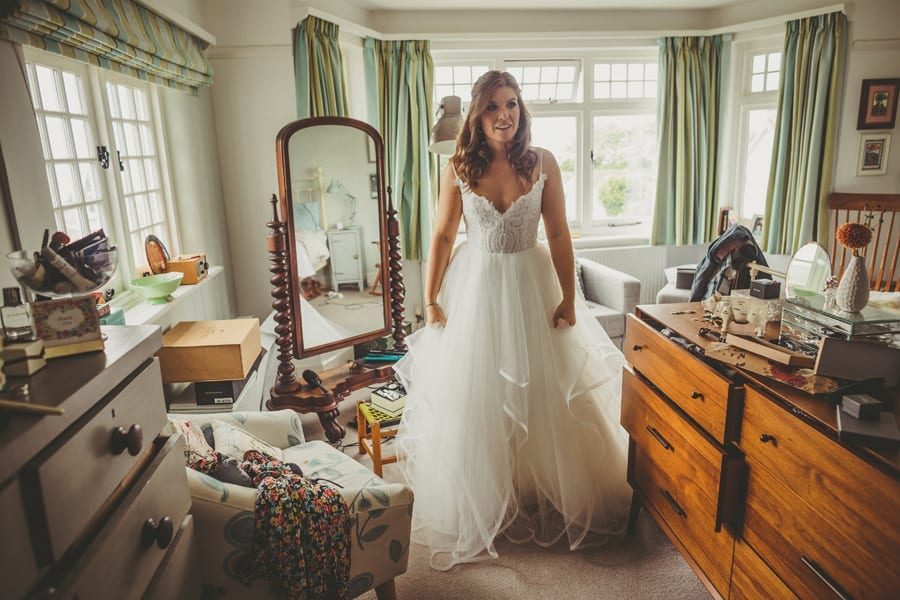 The bride stands in her wedding dress in her parents bedroom