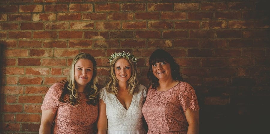 The bride and her bridesmaids pose for a photograph against a large brick wall at Over Barn