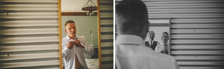 The groom fastens a button at the top of his shirt as he looks in the mirror