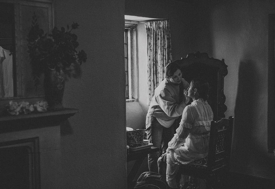 The makeup artist applies makeup onto the face of the bride next to a window in the cottage