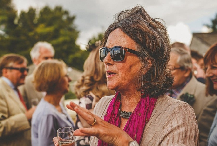 A wedding guest holding a flute of champagne with sunglasses on talks to a friend outside Silk Mill Studios