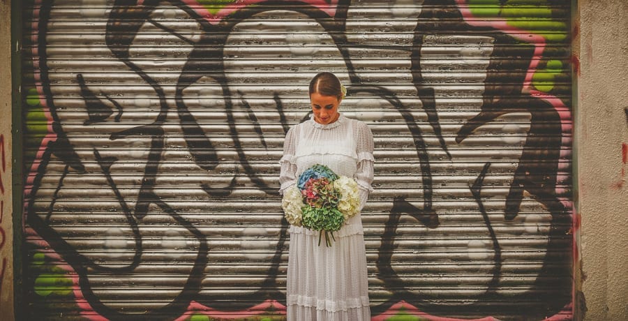 The bride looks down at her bouquet as she stands in front of a large metal shutter