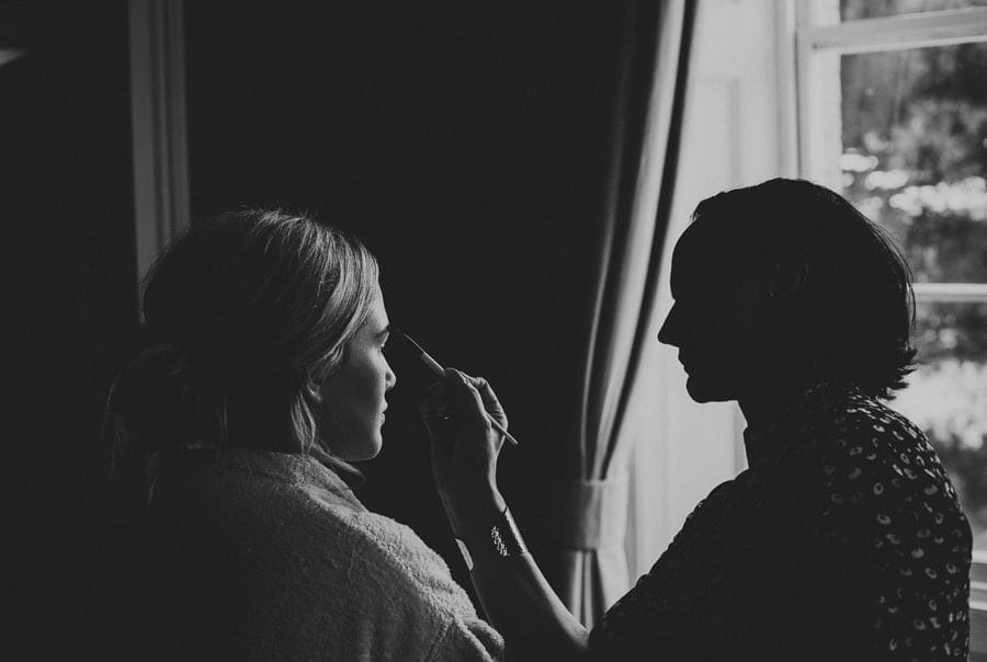 Bridesmaid sits on a chair next to the window while the makeup artist applies mascara