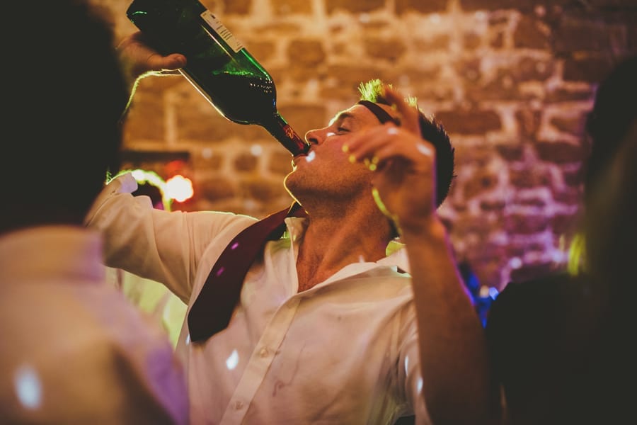 A wedding guest with a tie around his head drinks from a bottle of red wine