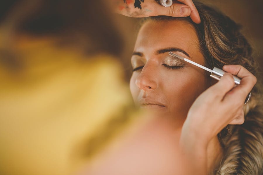A makeup artist carefully applies mascara onto the eyebrows of one of the bridesmaids in the cottage at Yurt retreat