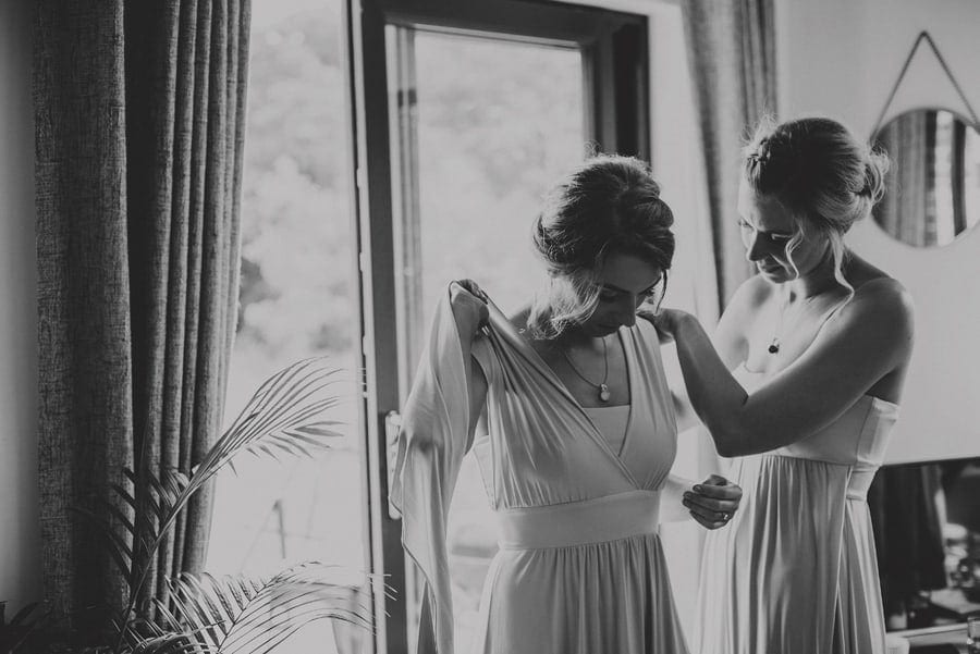 A Bridesmaid helps another bridesmaid to put on their dresses next to a window of the cottage