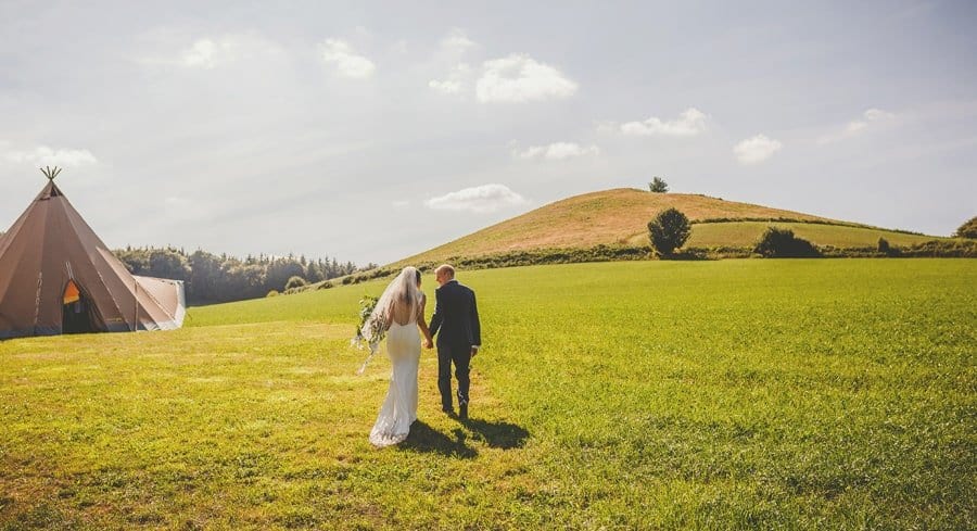 The bride and groom hold hands and walk towards the large tipi in the field at yurt retreat