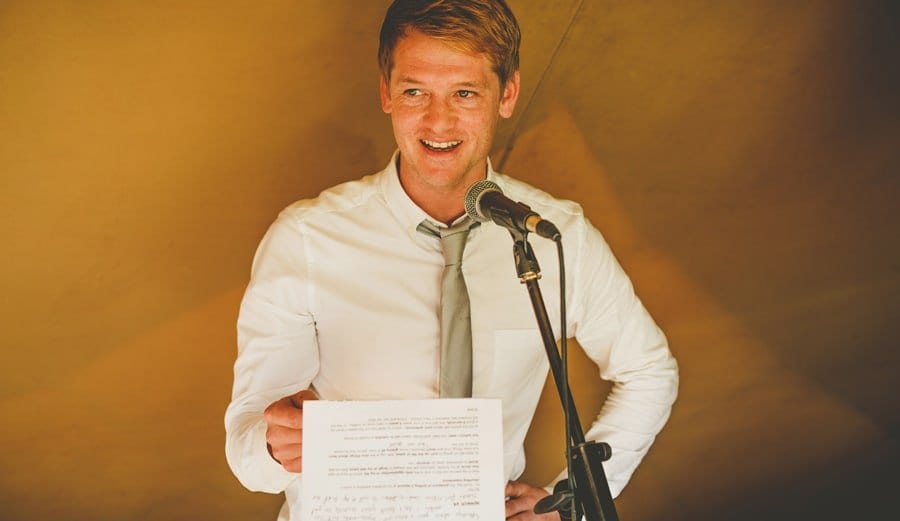 The best man holds a piece of paper in his hand and delivers his speech to the wedding guests in the tipi
