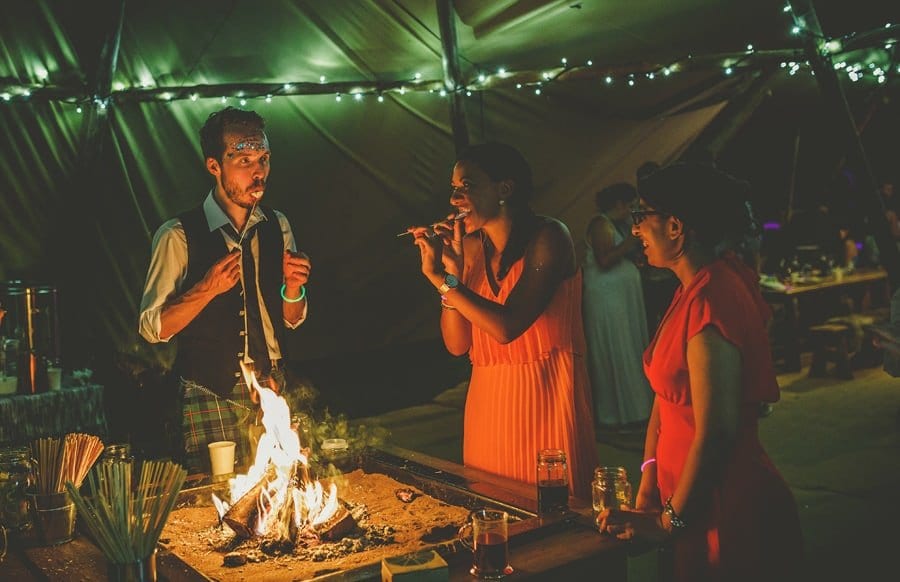 Wedding guests toast marshmellows over an open lit fire in the tipi