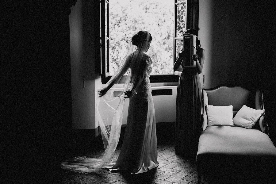 A bridesmaid holds a mirror up as the bride looks at herself as she stands next to a large window