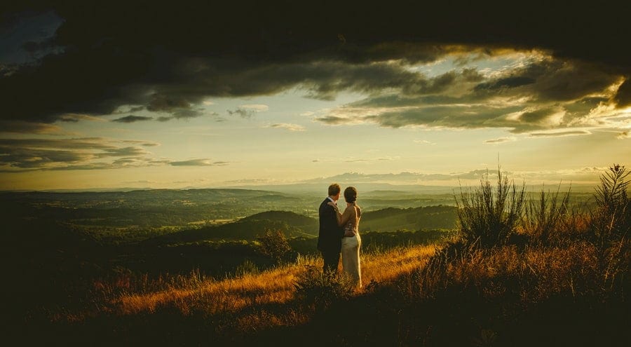 The bride and groom stand next to each other on top of a hill and look towards the distant hills as the sun goes down