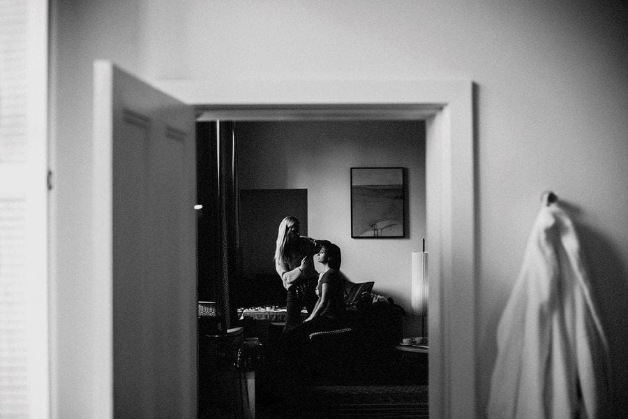 A makeup artist applies make up to the face of the bride who is sat next to a large window in a hotel room